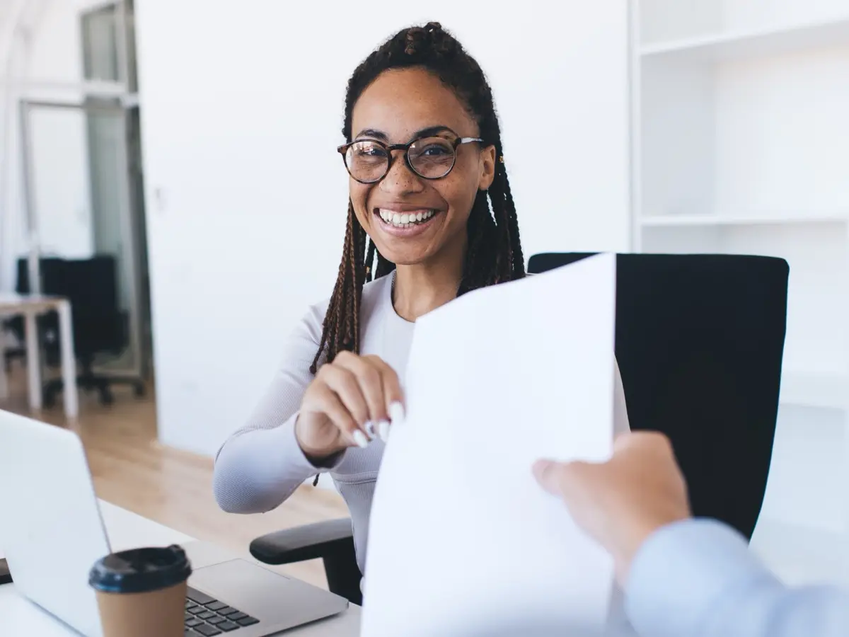 Smiling woman at a desk handing a printed flyer to a visitor